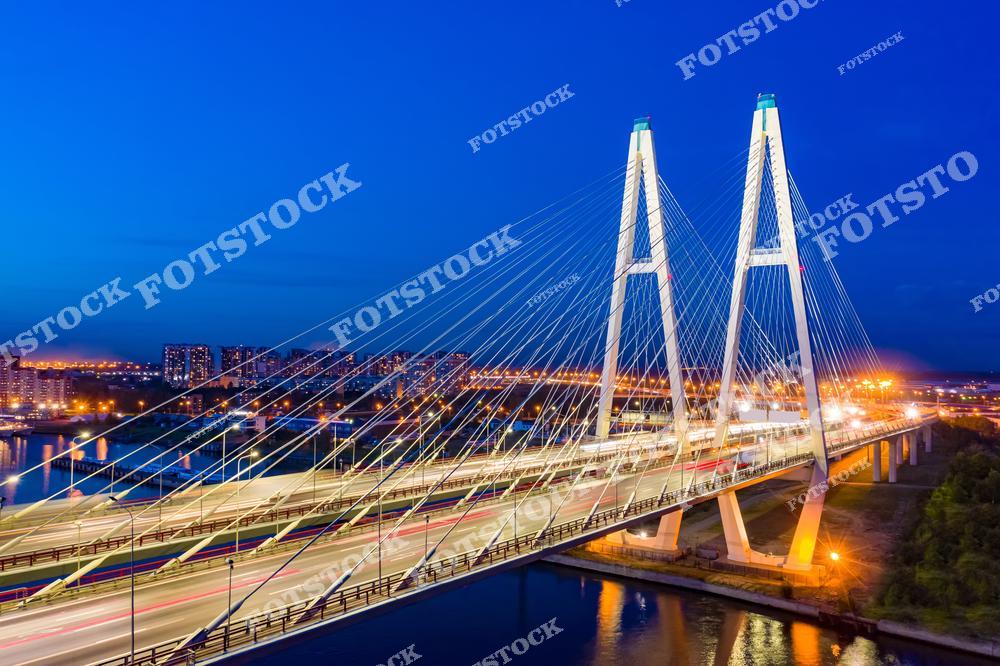 На снимке изображён Большой Обуховский мост — уникальный вантовый мост через Неву и один из символов современной инфраструктуры Санкт-Петербурга. Мост освещён яркой иллюминацией, подчёркивающей его конструктивную элегантность. На фоне видны жилые районы города и огни магистралей. Этот мост является важным элементом транспортной системы города. Снимок выполнен в тёмное время суток с высоты, что усиливает эффект движения и урбанистики.